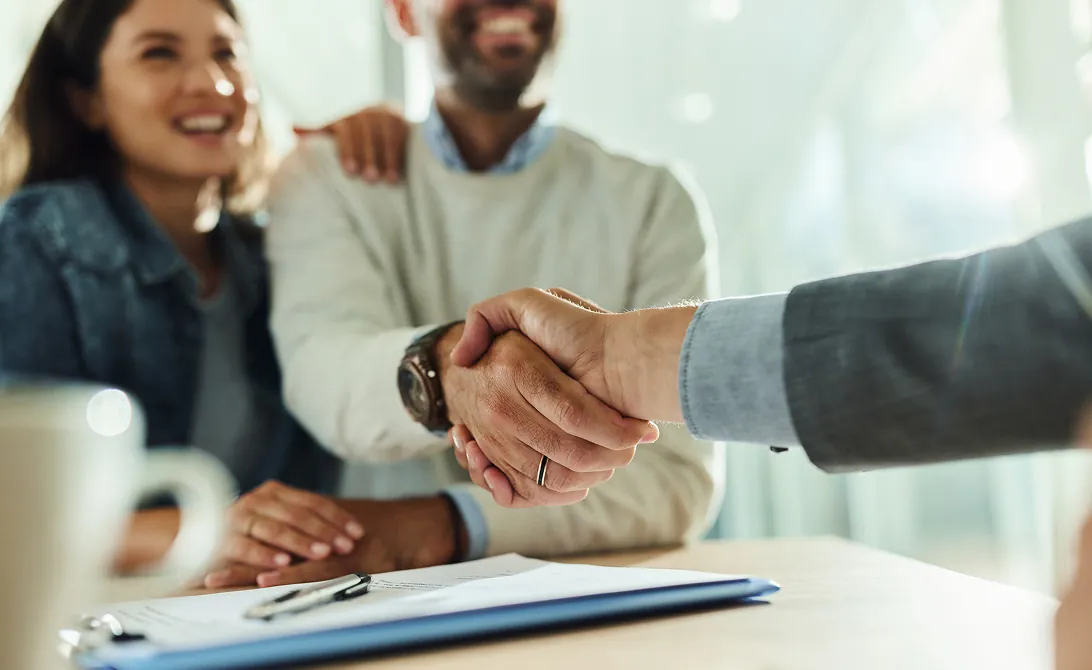 A smiling couple celebrates while someone shakes hands across a table with documents. The scene conveys success and agreement in a professional setting.
