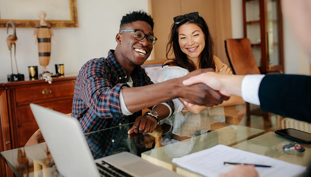 A smiling couple sits at a glass table, shaking hands with someone off-camera. Papers and a laptop are on the table, suggesting a professional meeting.